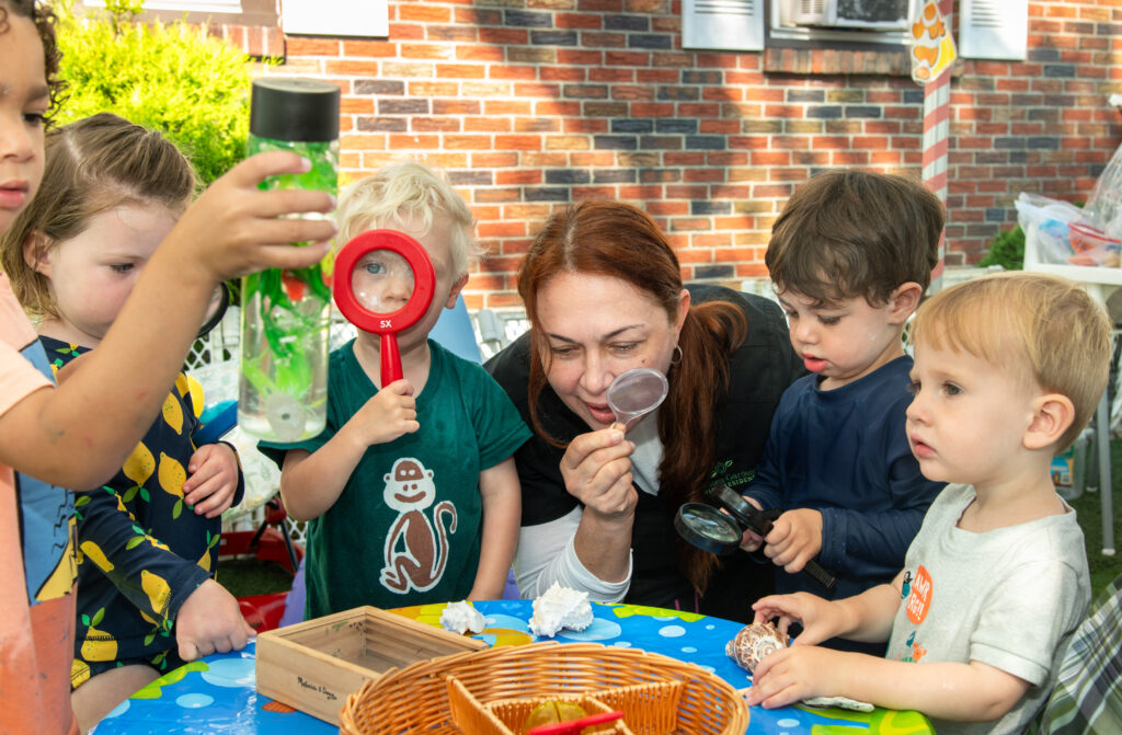 An early childhood educator is showing a group of children how to use a magnifying glass by holding it up to her eye and looking through it at objects on a table. 