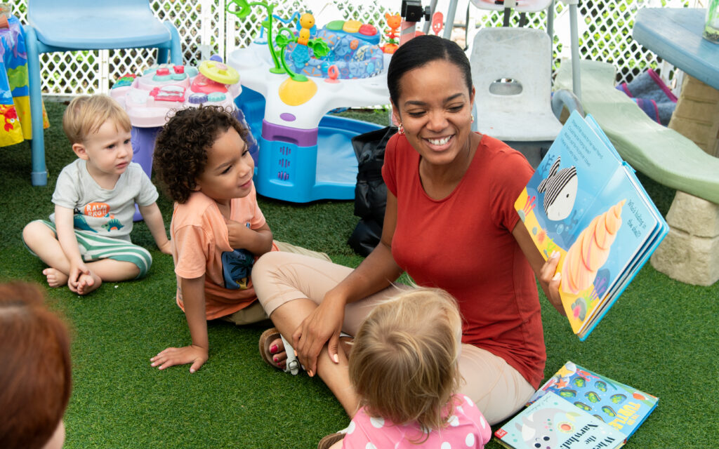 An early childhood educator is sitting cross legged on the grass holding up a book to show the pictures to a few children seated on either side of the educator. 