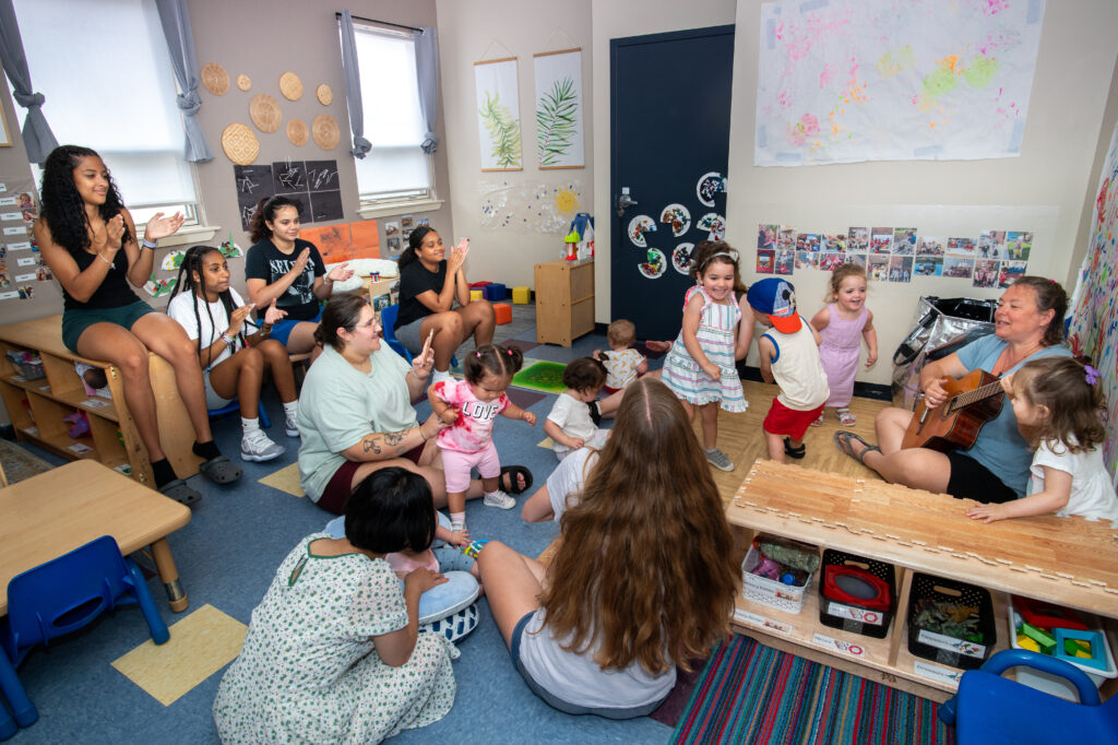 A classroom of children and educators clapping, dancing, and singing while a person plays the guitar. 