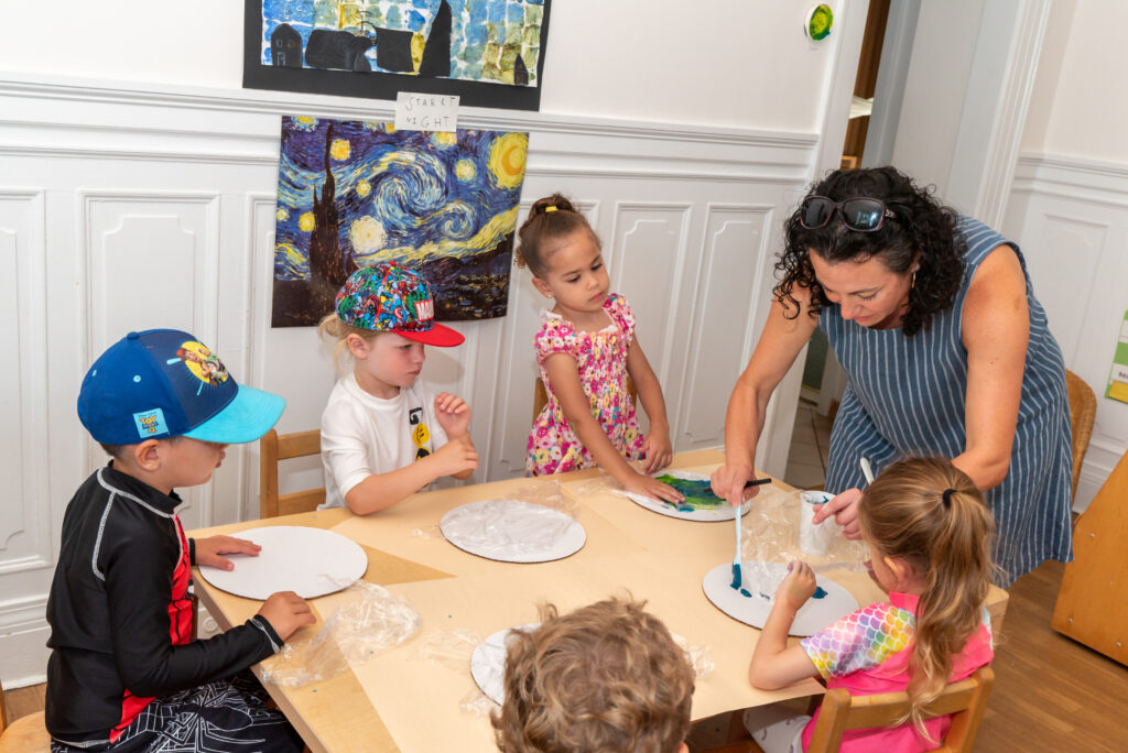 An educator leans over a table working with a group of children to demonstrate how to paint